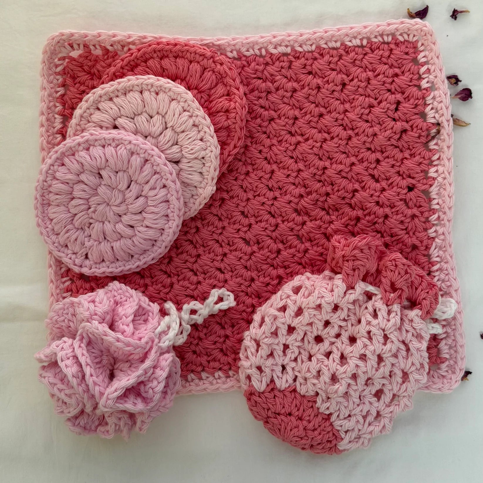 Handmade crochet bath set with pink, rose, and white cotton yarn poufs and face scrubbies arranged on neutral background